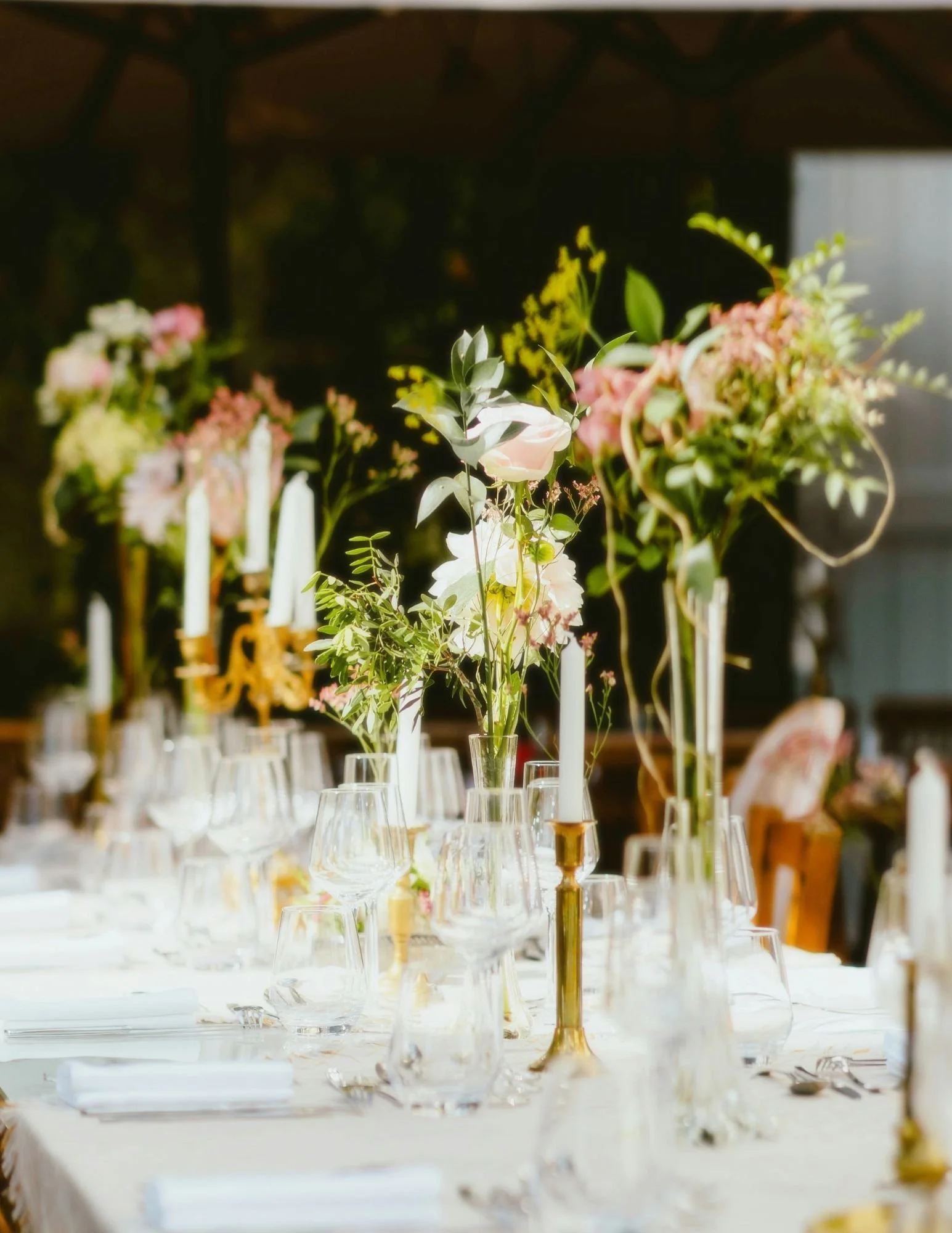 Table with soft light and clear glass stemware. Brass candle sticks and candelabra with soft white candlesticks. Tall etherial flower arrangements with whimsical greenery and pastel colored florals.