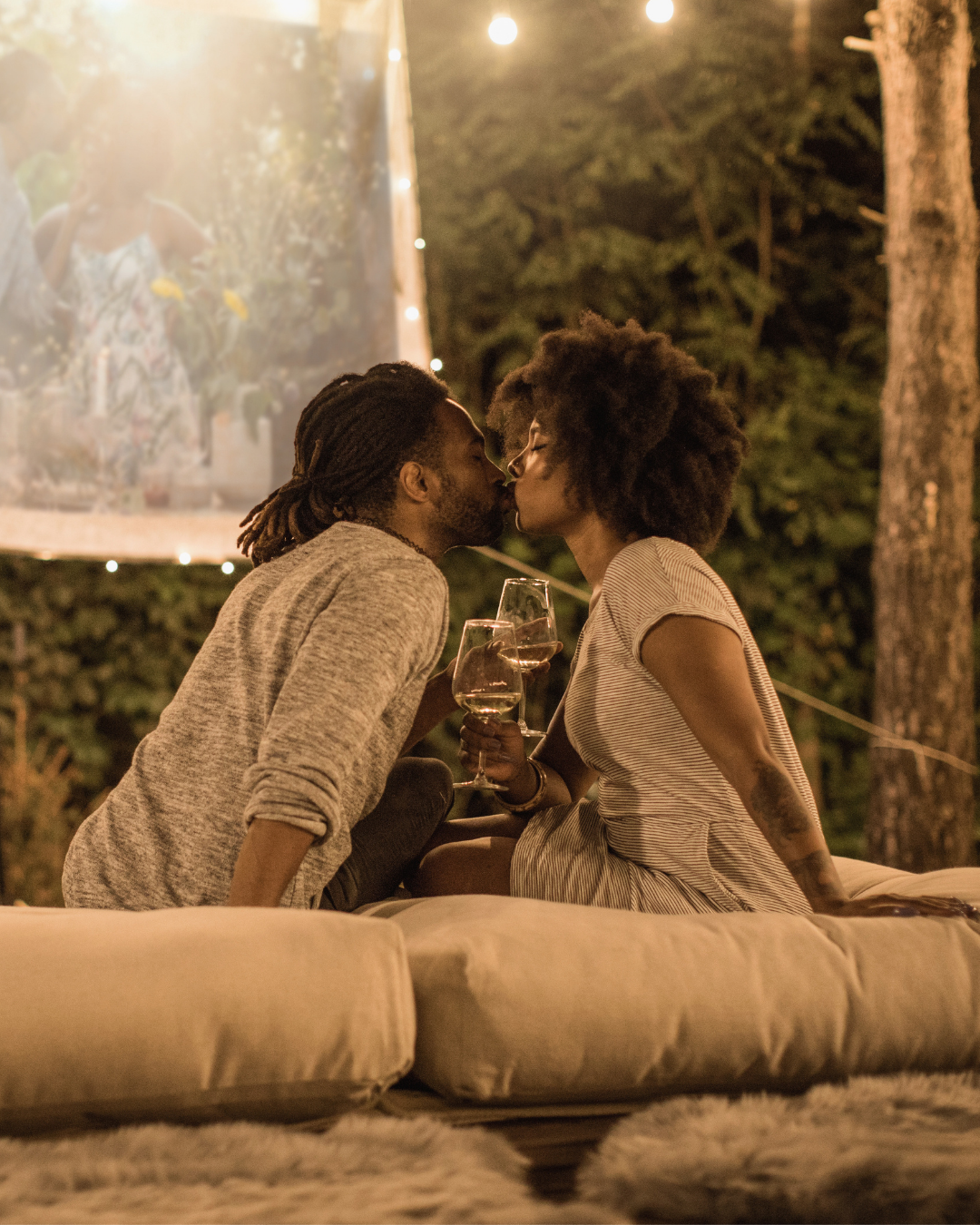 A couple kissing at an outdoor evening gathering, sitting on a cushioned surface, holding wine glasses, surrounded by string lights and trees.