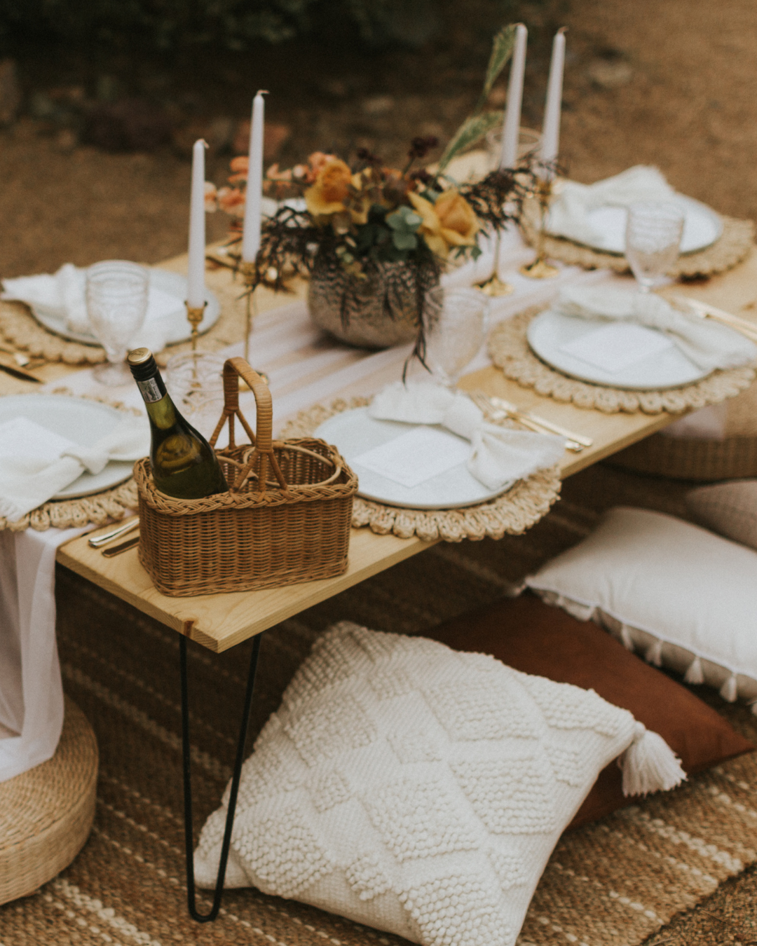A rustic outdoor dining table set with white plates, napkins, gold utensils, candles, a floral centerpiece, a basket with a bottle of wine, and pillows on a woven rug.