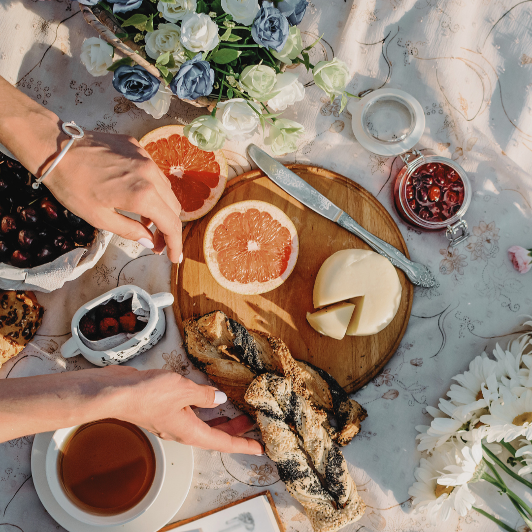 Picnic table setting with brunch items including sliced grapefruit, cheese, pastries, tea, fruit and flowers