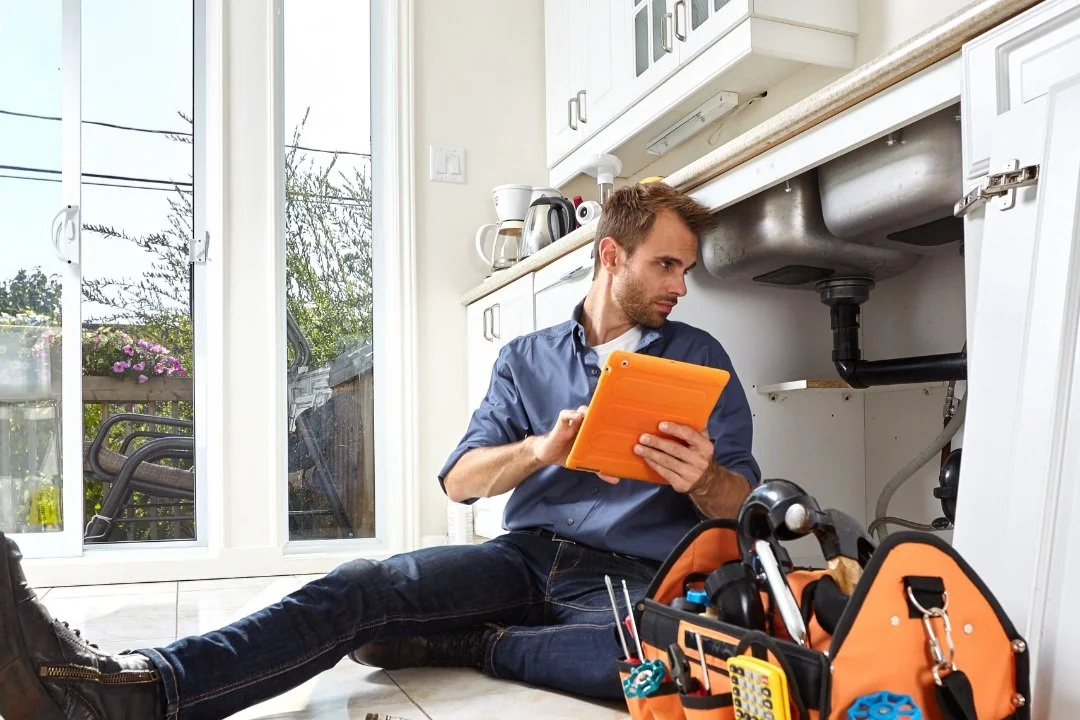 A man working under a kitchen sink with plumbing issues, holding a tablet, surrounded by tools in a tool bag.