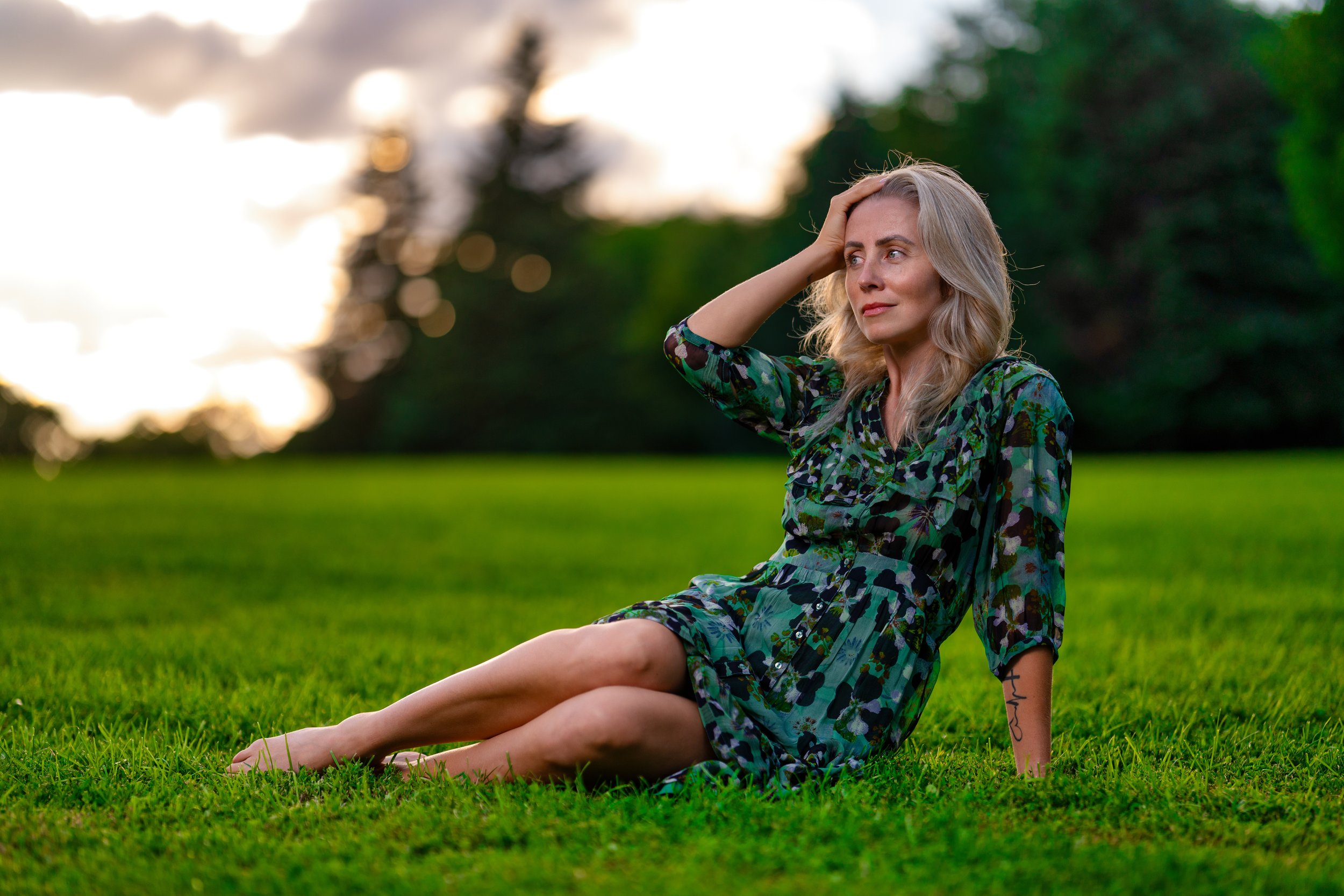 A woman sitting on the grass in a park during sunset, wearing a patterned dress, with one hand on her head, looking contemplative.