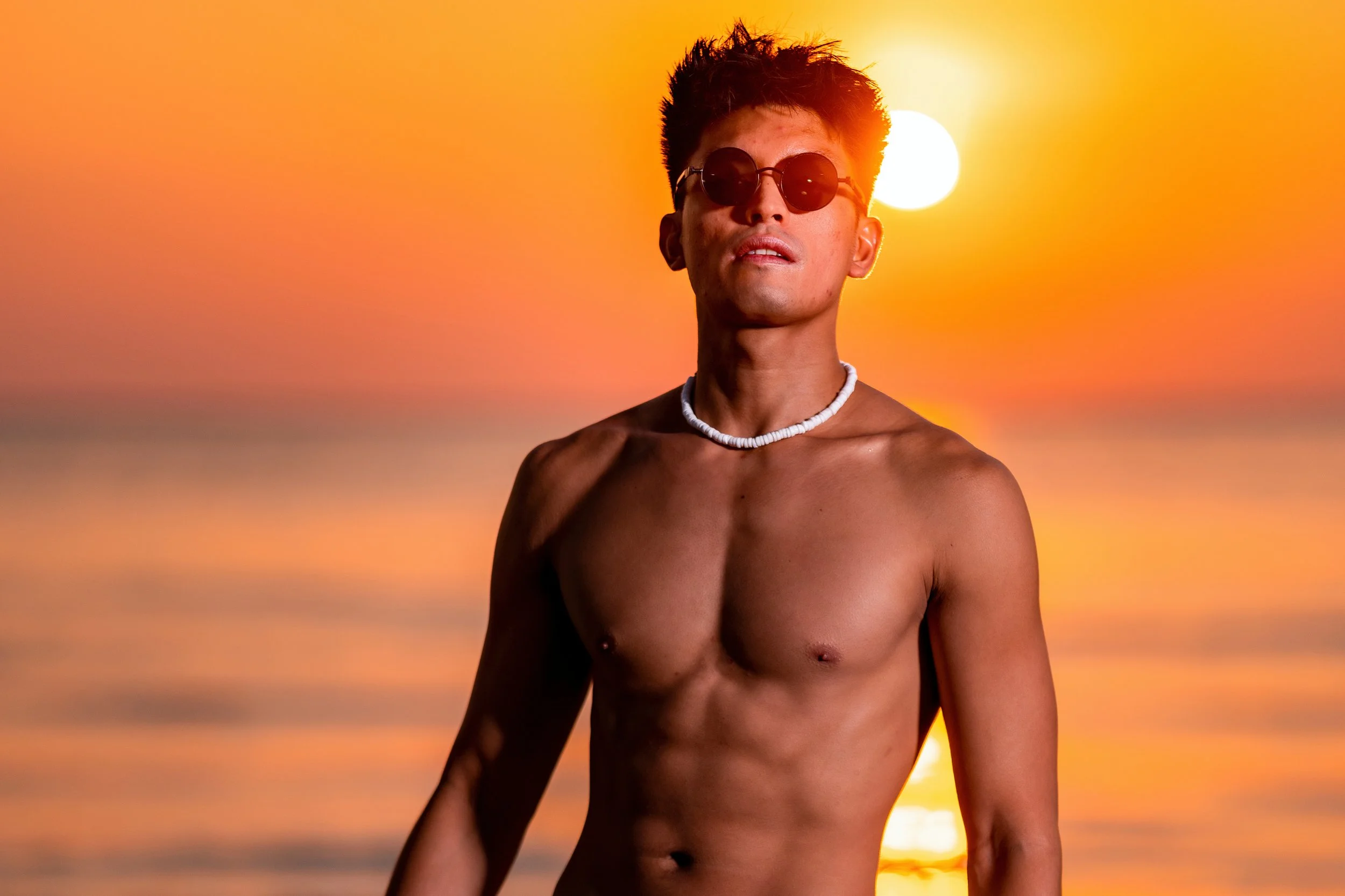 A shirtless young man with dark hair wearing sunglasses and a white shell necklace, standing in front of a sunset over the ocean with the sun in the background.