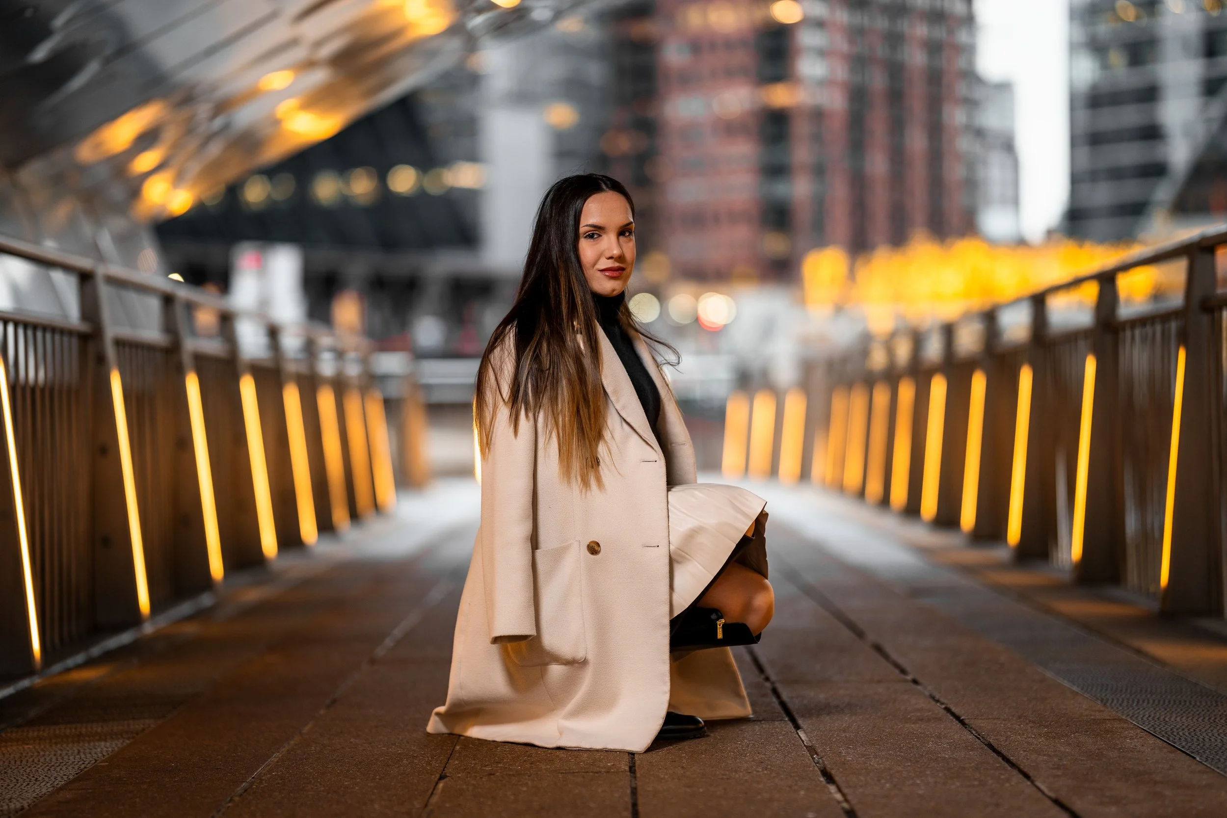 A woman with long dark hair in a beige coat kneeling on a city bridge at sunset, with a blurred urban background and glowing yellow lights along the bridge railing.