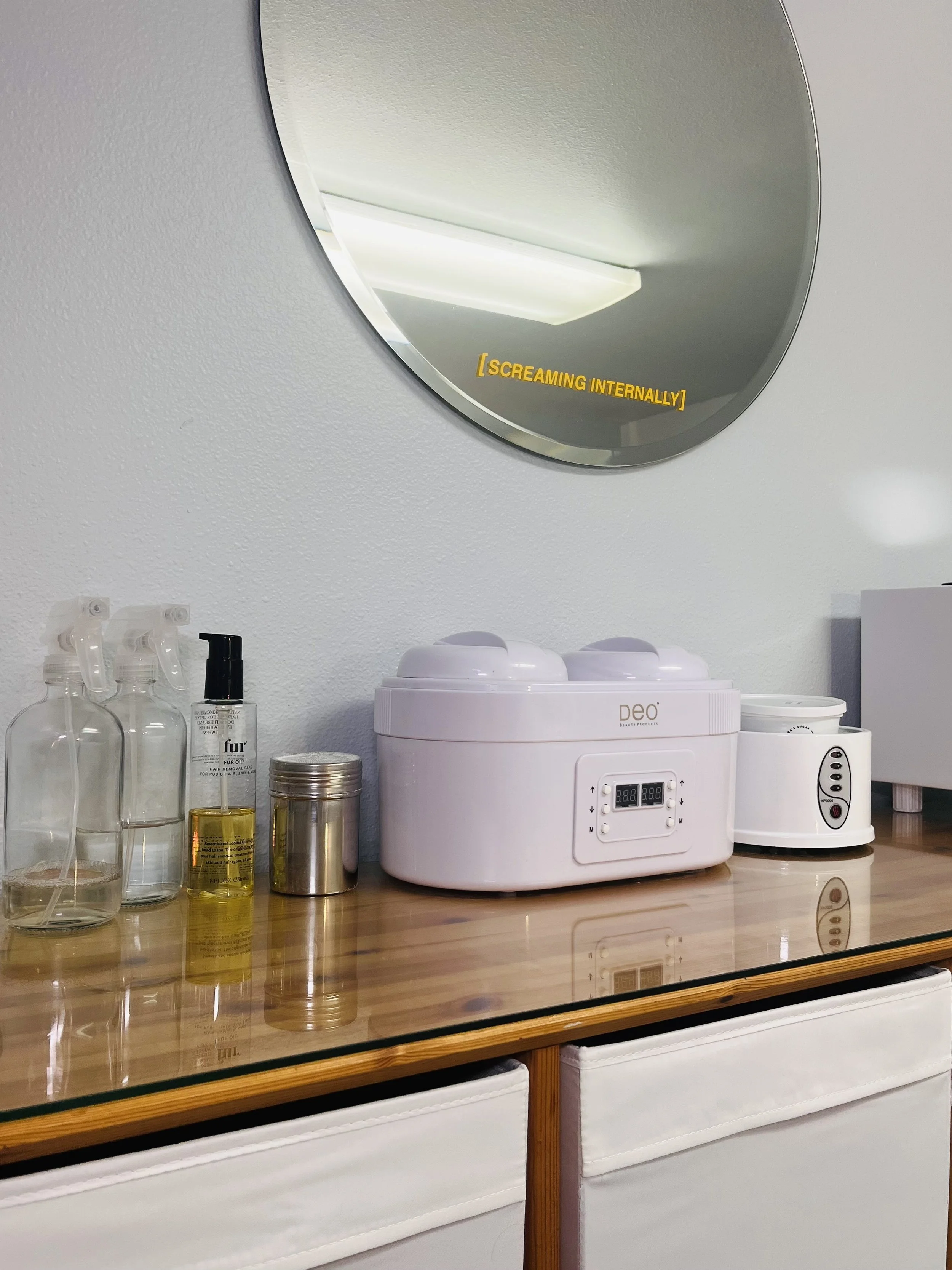 A wooden countertop with various bottles, a small jar, and a white steam treatment machine. A round mirror above reflects a ceiling light and has the text '[SCREAMING INTERNALLY]' on it.