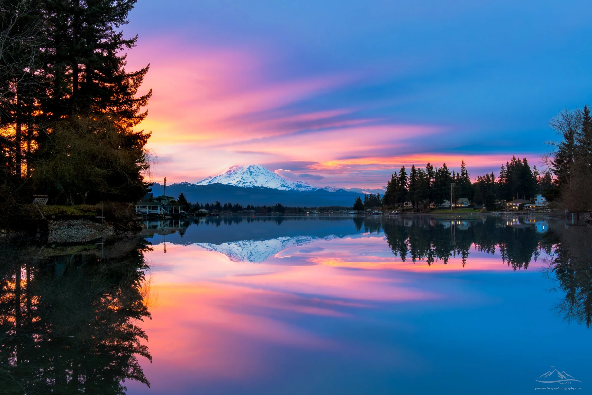 A serene landscape featuring Lake Tapps reflecting a colorful sunset with pink, purple, and orange hues. In the distance, snow-capped Mt. Rainier is visible against a partly cloudy sky, with trees and houses along the shoreline. 