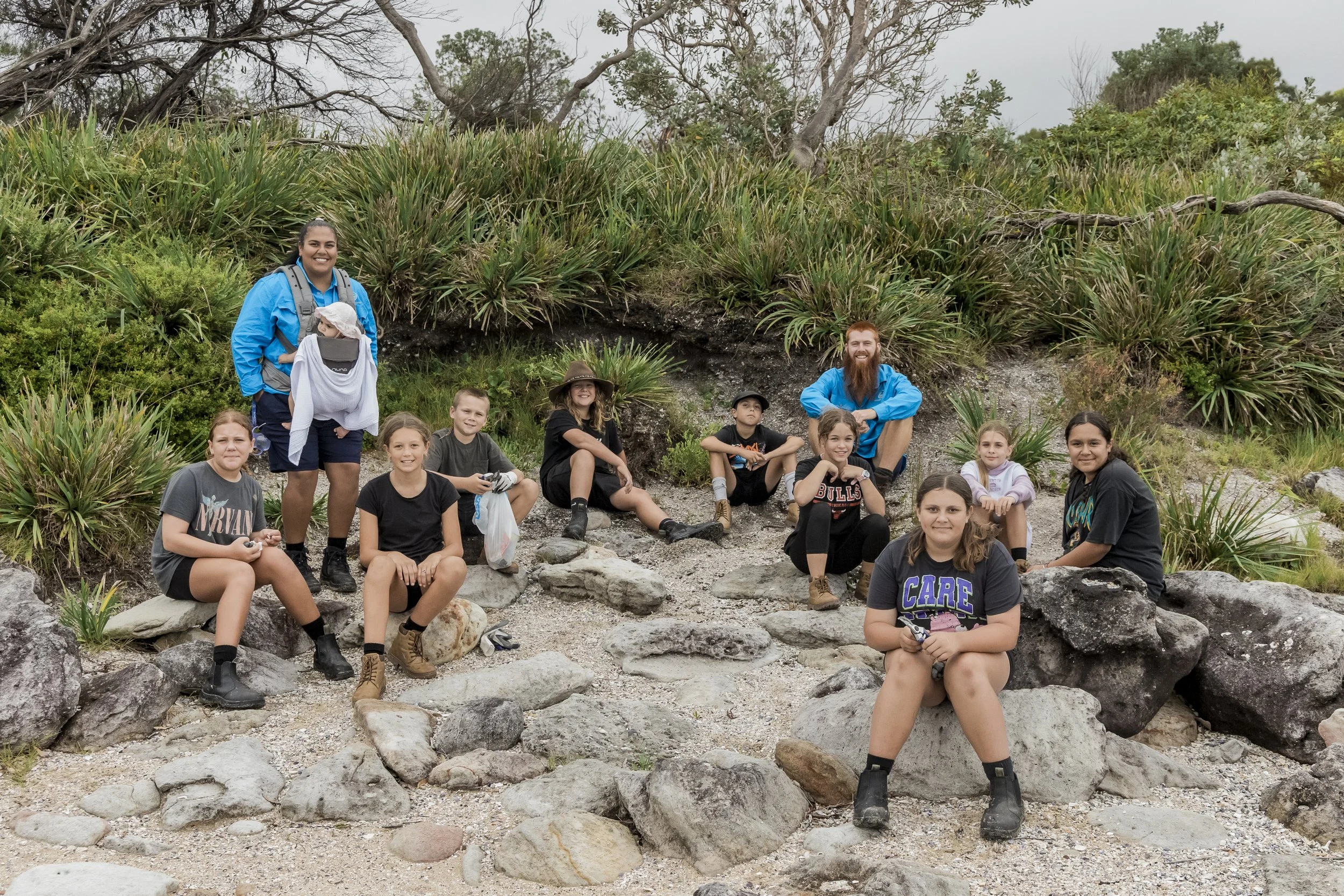 A group of ten people, including children and adults, off a hike in a natural area with rocks and greenery. They are sitting or kneeling on large rocks, with two adults at the back and children in front, all smiling for the photo.