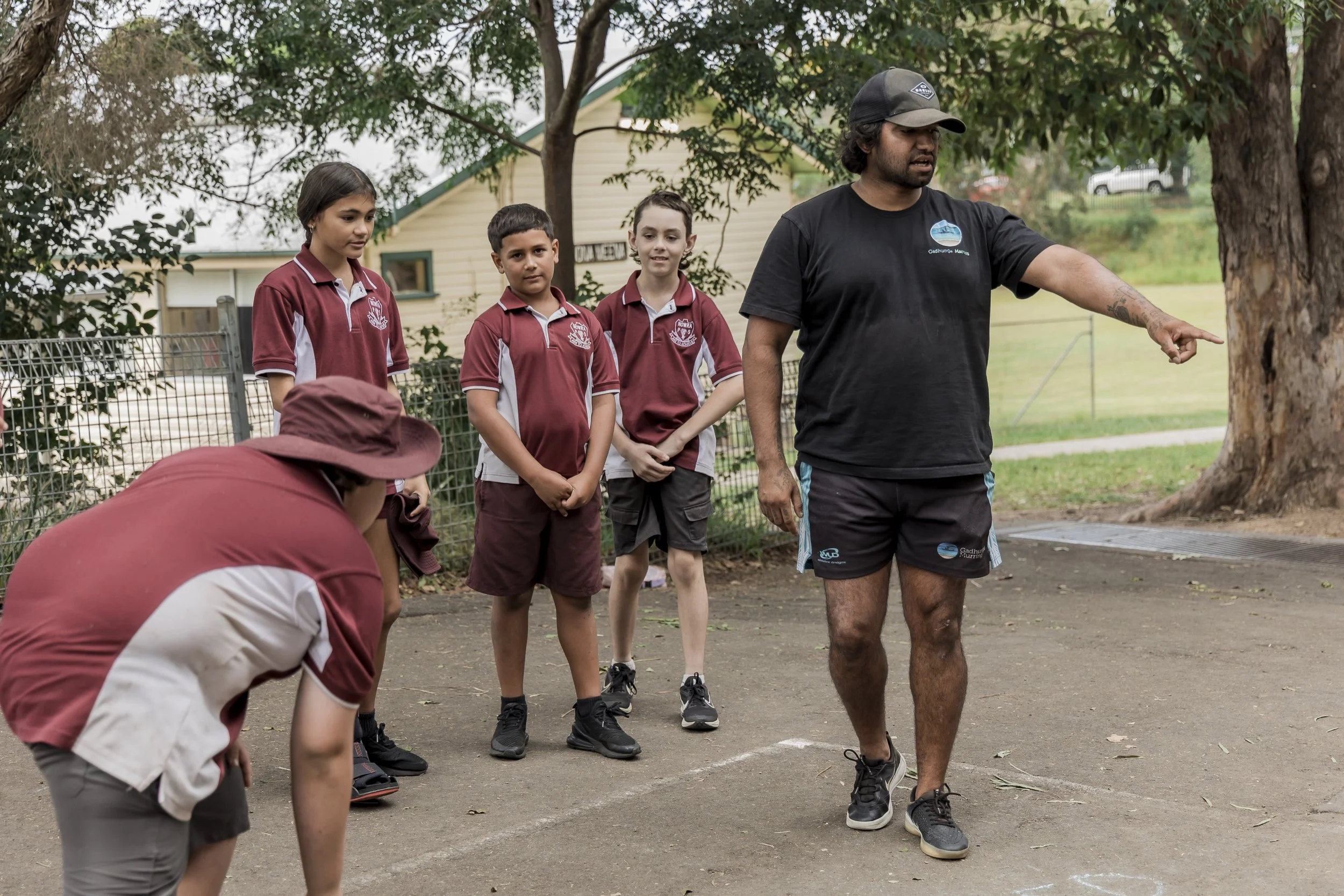 Outdoor scene with four children in maroon uniforms, a man in black explaining something, and another person in a maroon hat and shirt crouched down, near a tree and fence.