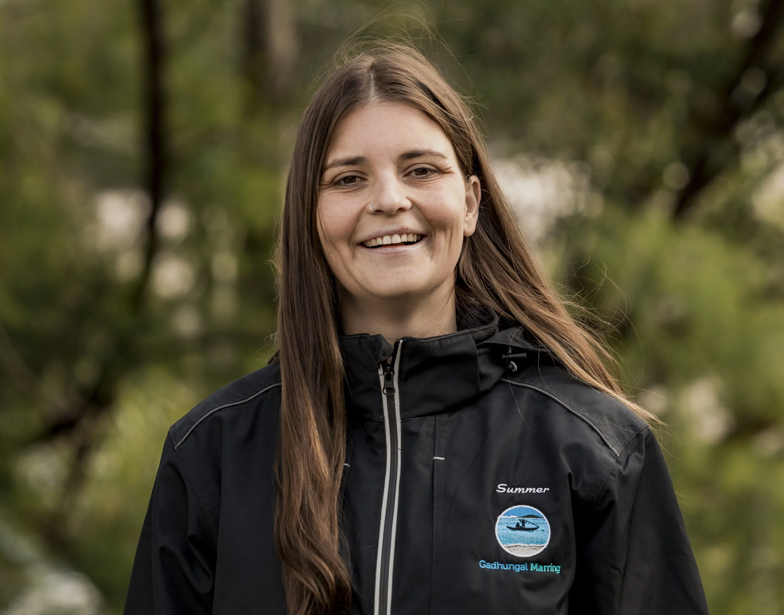 A young woman with long brown hair smiling outdoors, wearing a black jacket with patches that read 'Summer' and 'Gadhungul Marring', and a background of green trees.