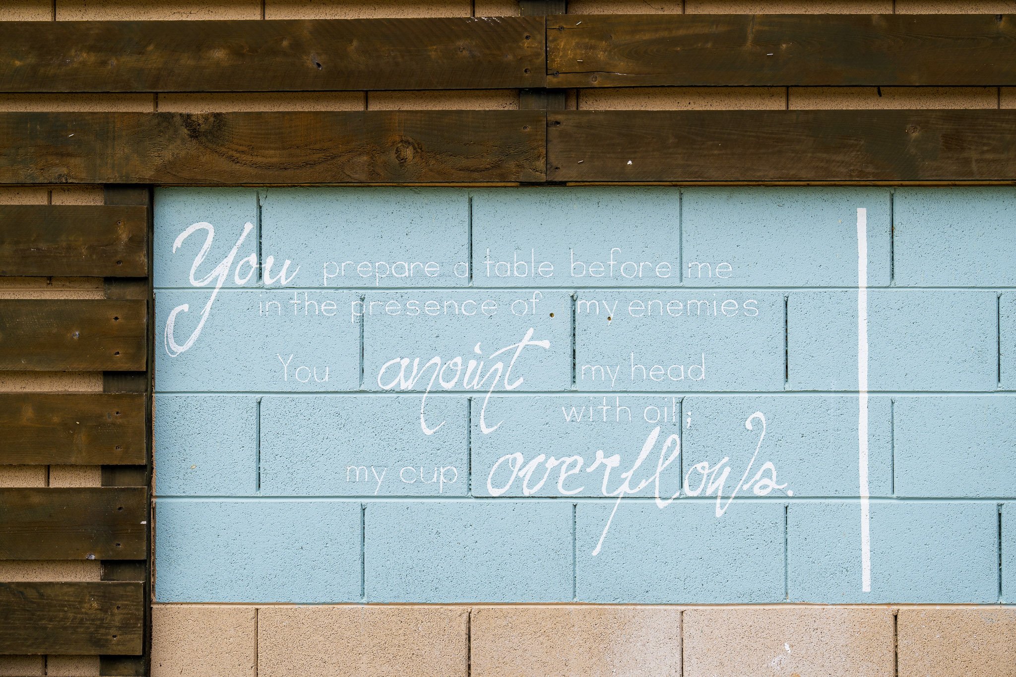 Bible verse on a blue brick wall with wooden framing, featuring Psalm 23:5 about preparing a table and anointing with oil.