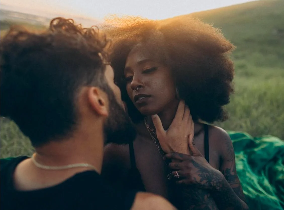 A man and woman with dark curly hair in an intimate outdoor moment at sunset, close-up, with grass in the background.