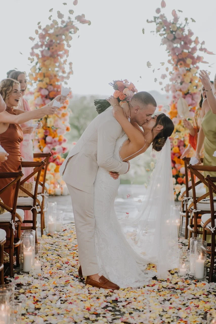 Bride and groom sharing a romantic kiss under a floral arch, surrounded by cheering guests and flower petals.
