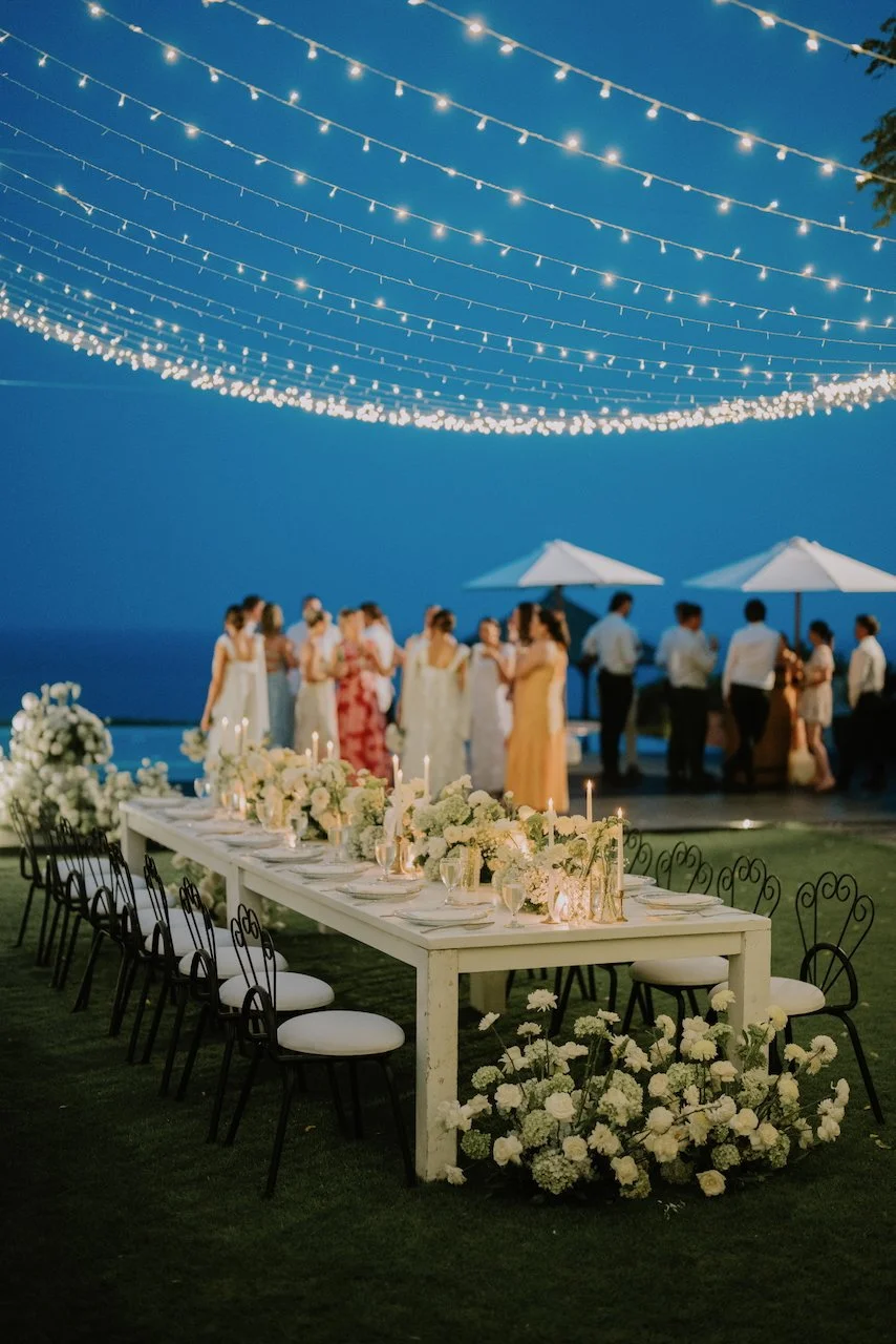 Romantic Bali wedding reception with ocean view, elegant white floral table decorations, glowing candles, and string lights under the evening sky