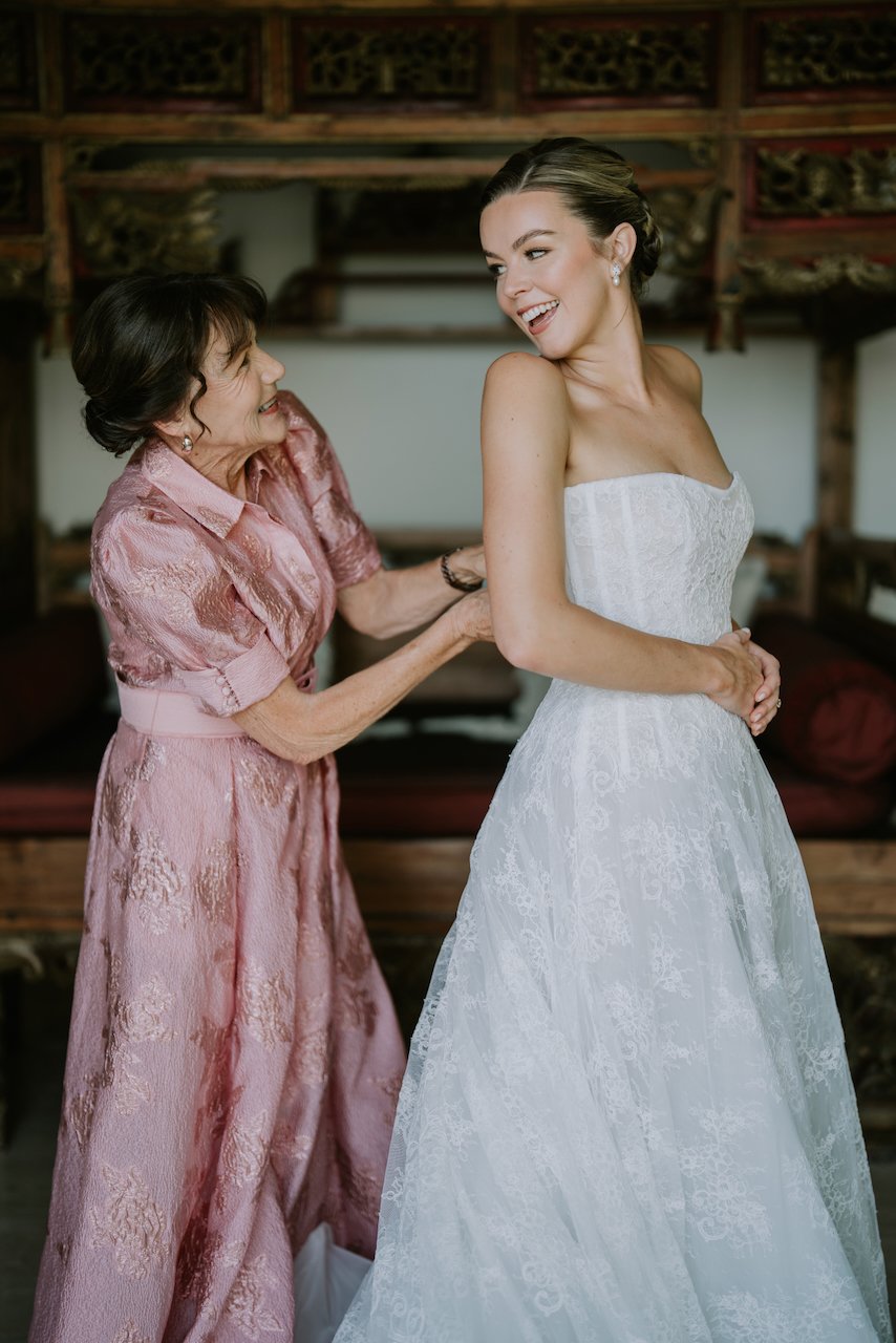 Mother helping bride into lace wedding gown before Bali villa ceremony.