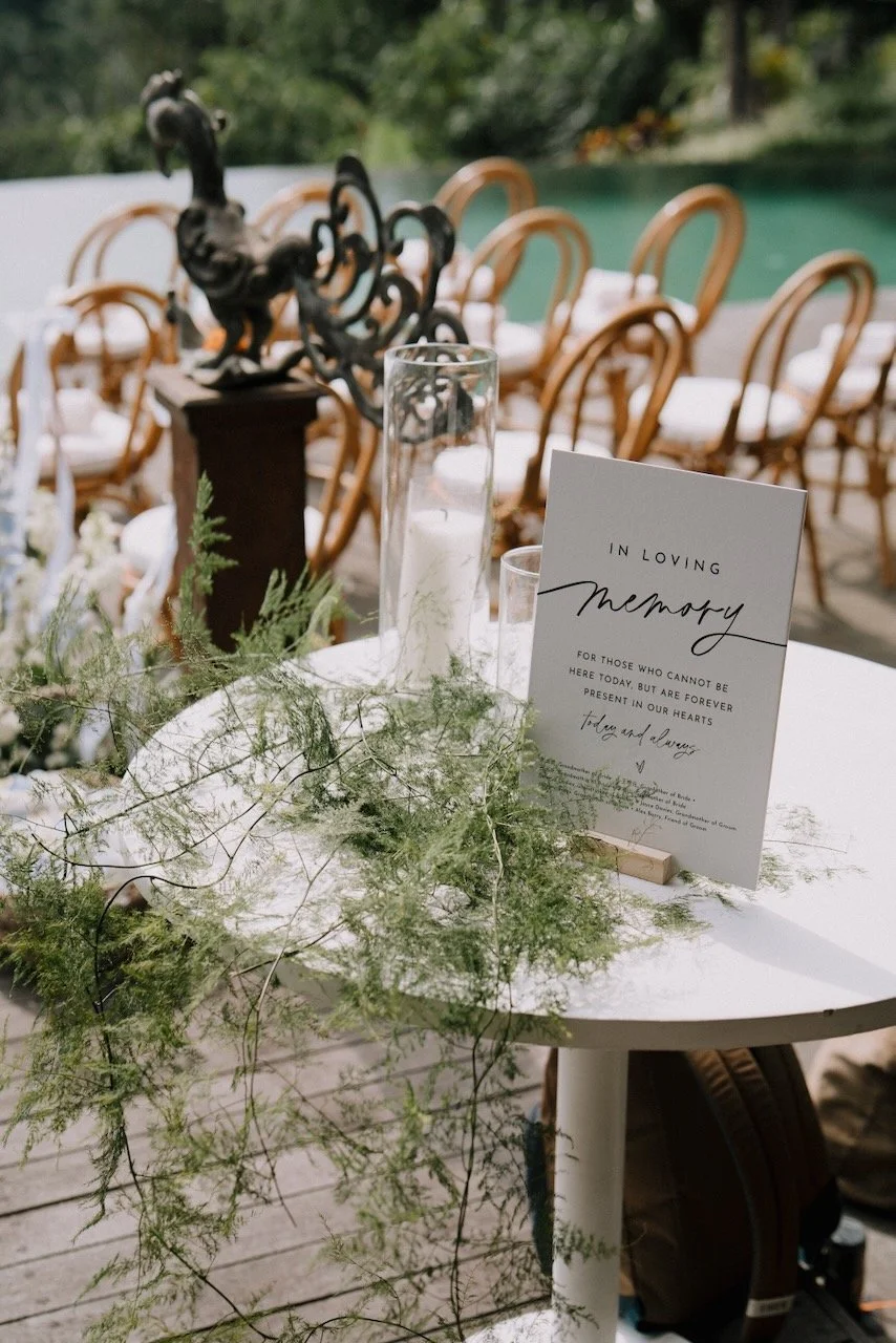 Bali wedding memorial table with candle and “In Loving Memory” sign overlooking tropical poolside ceremony setup.