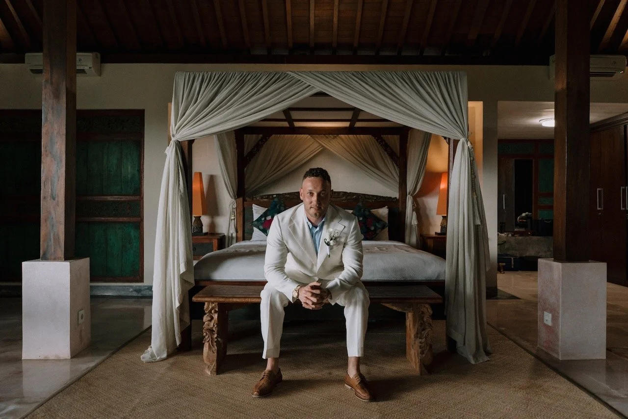Groom in white suit sitting in Bali villa bedroom before wedding ceremony, calm and thoughtful.