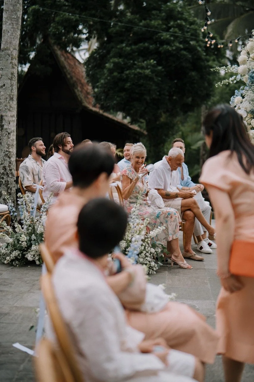 Wedding guests seated among floral arrangements at outdoor Bali ceremony, smiling and chatting.