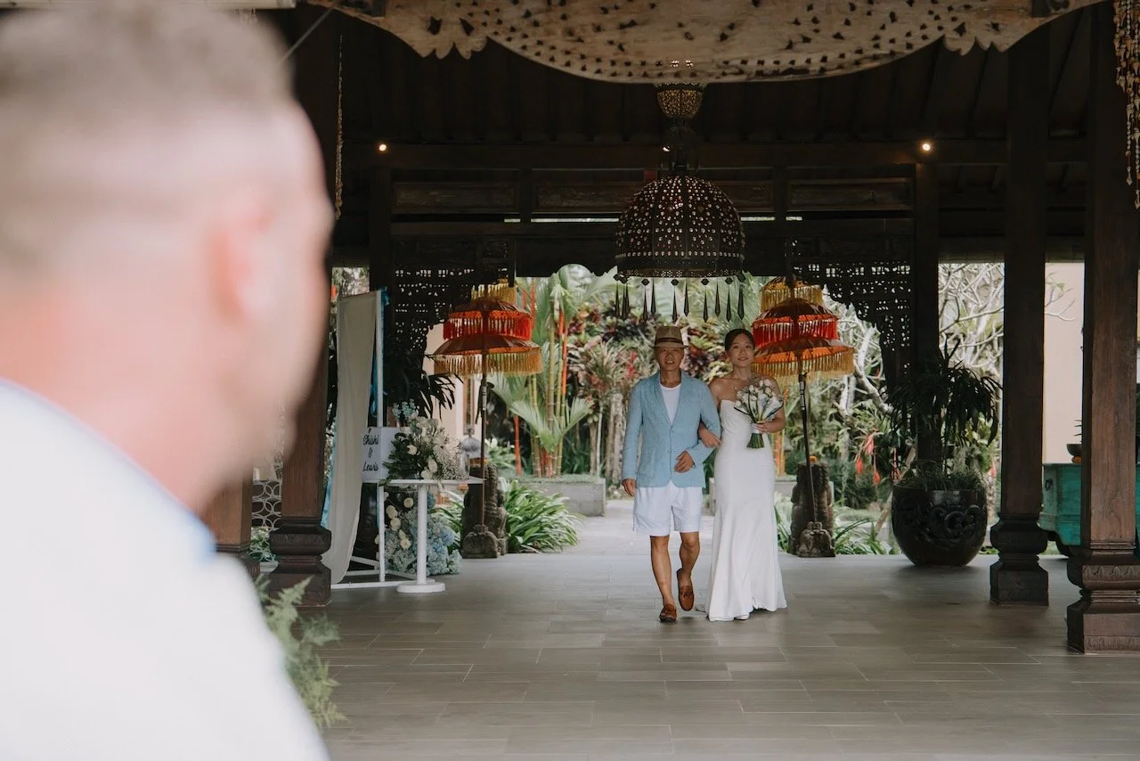 Bride walking down the aisle arm-in-arm with family member at Bali ceremony entrance decorated with lanterns.