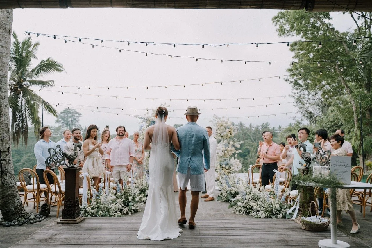 Bride walking towards groom down aisle adorned with white flowers and string lights at outdoor Bali wedding overlooking jungle.