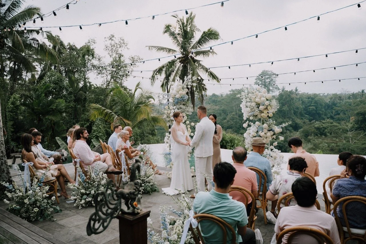 Outdoor Bali wedding ceremony with couple exchanging vows overlooking tropical jungle and infinity pool under string lights.