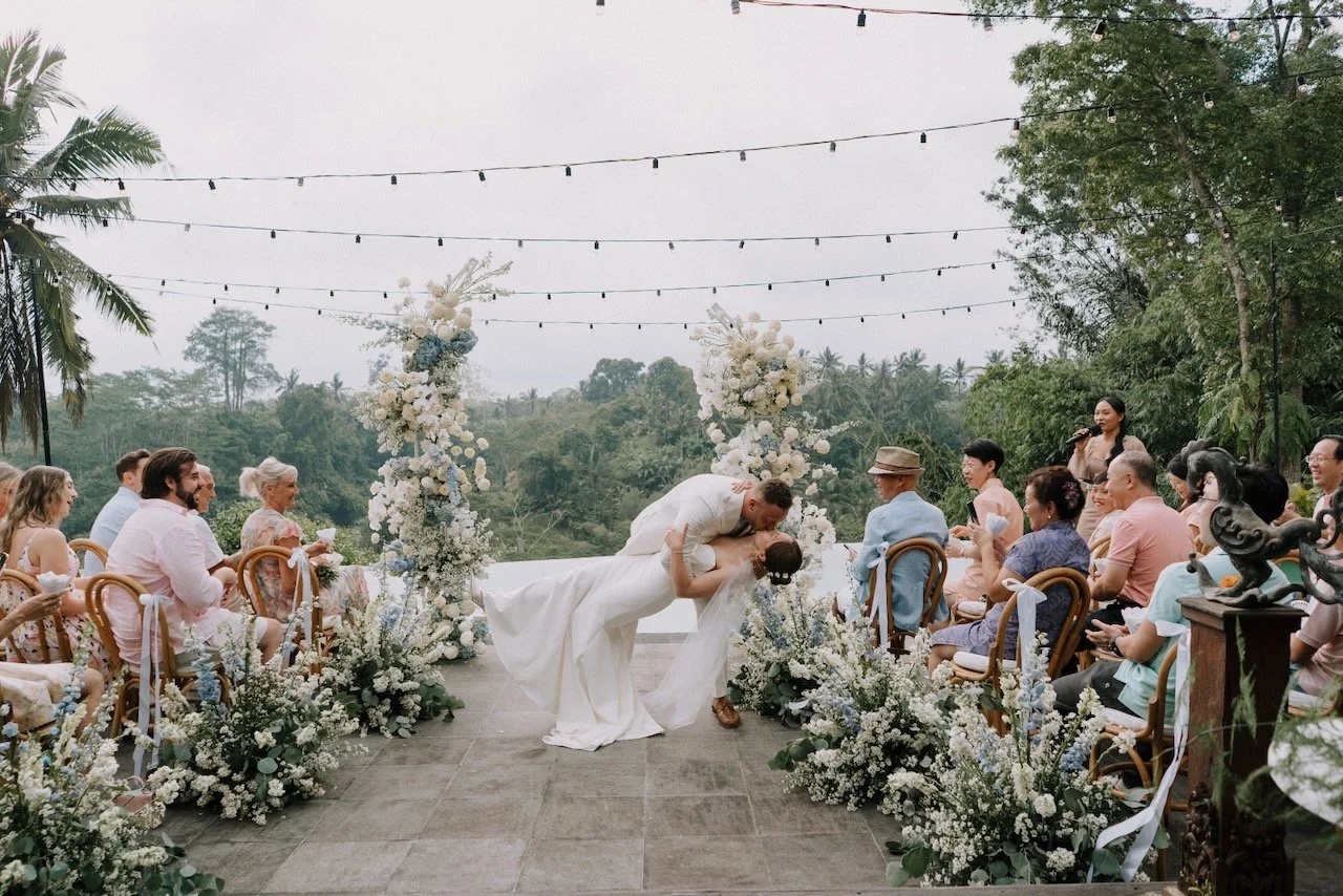 Groom dipping bride for a romantic kiss during Bali wedding ceremony surrounded by lush florals and cheering guests.