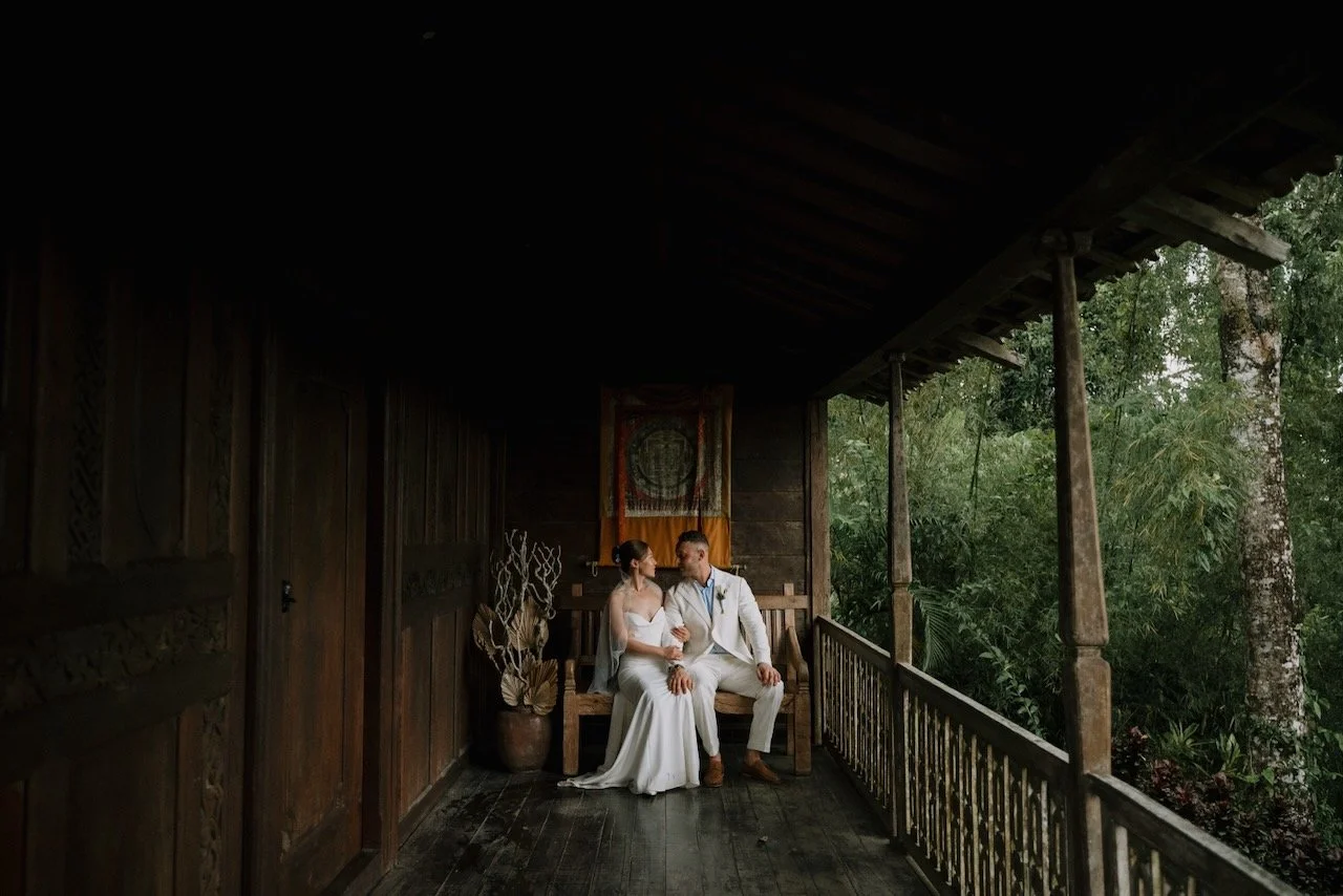 Bride and groom sitting together on wooden bench in traditional Balinese pavilion, sharing a quiet intimate moment.