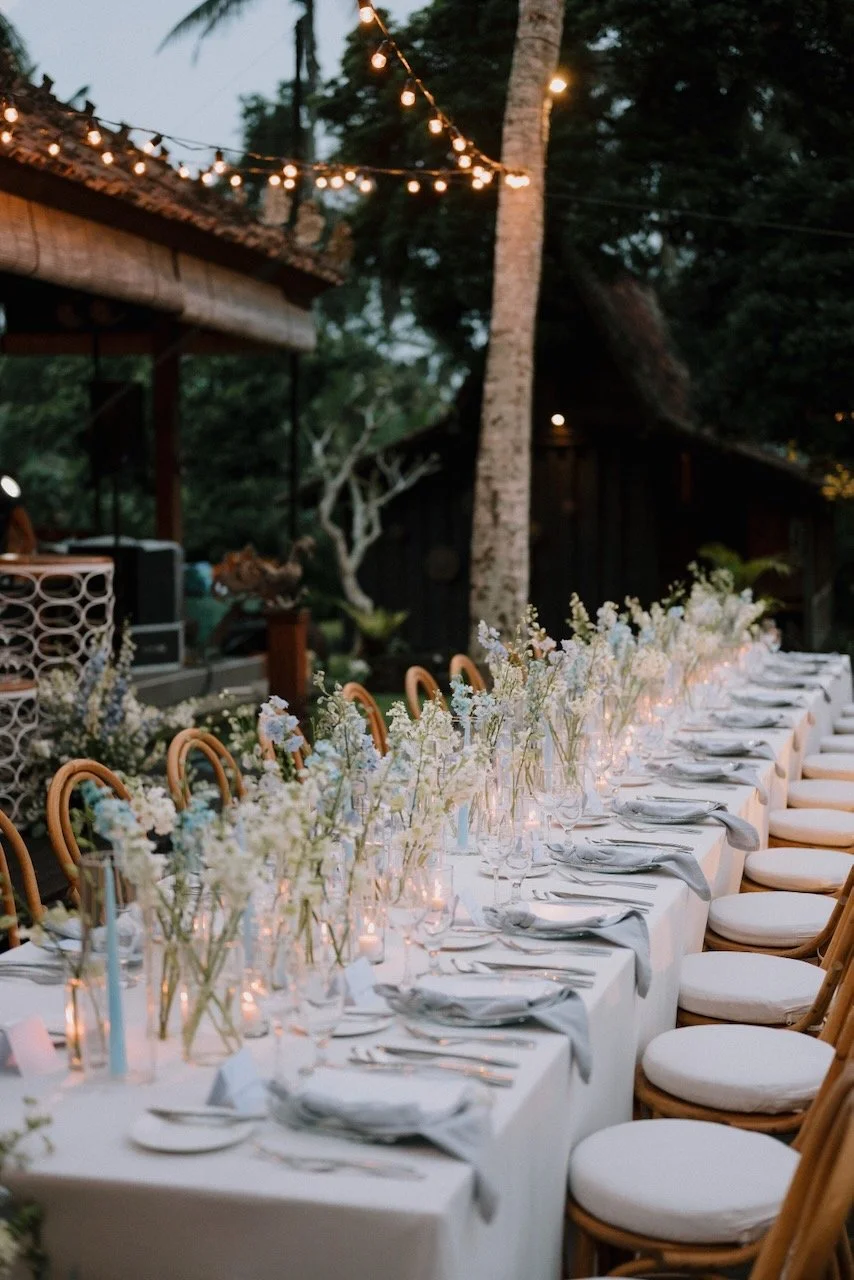 Elegant long reception table with white linens, pastel flowers and candlelight at outdoor Bali wedding dinner under string lights.