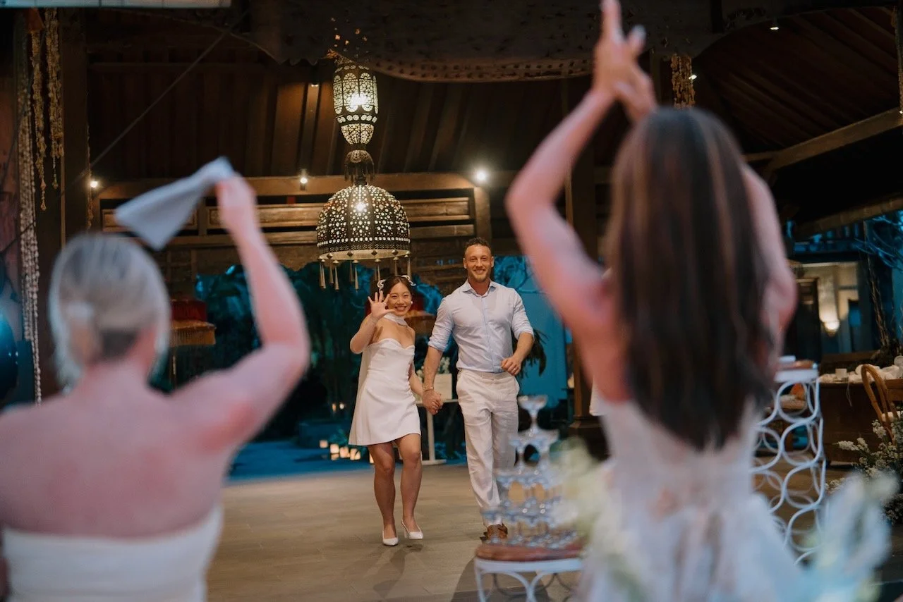 Bride and groom entering wedding reception in Bali, greeted by cheering guests and festive lantern lights.