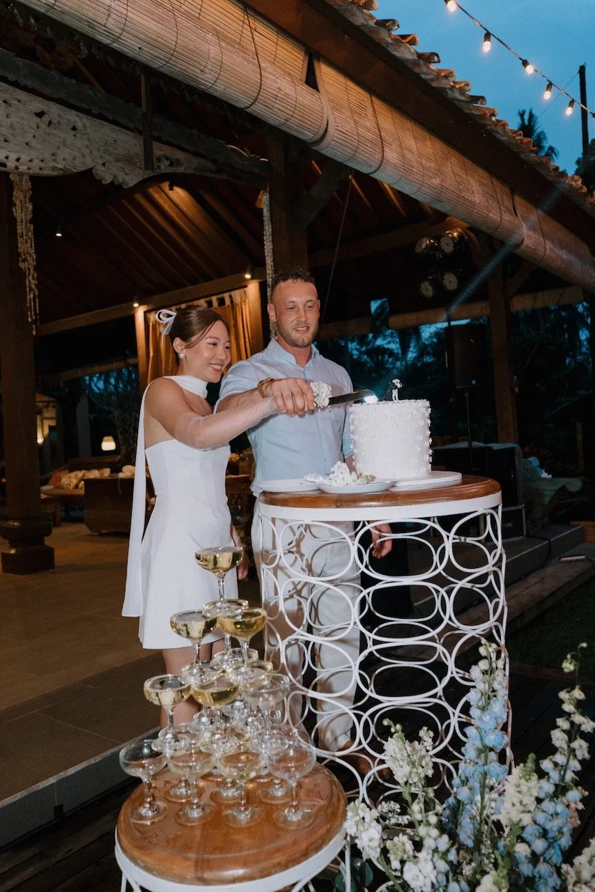 Bride and groom cutting a white textured wedding cake beside champagne tower during Bali wedding reception.