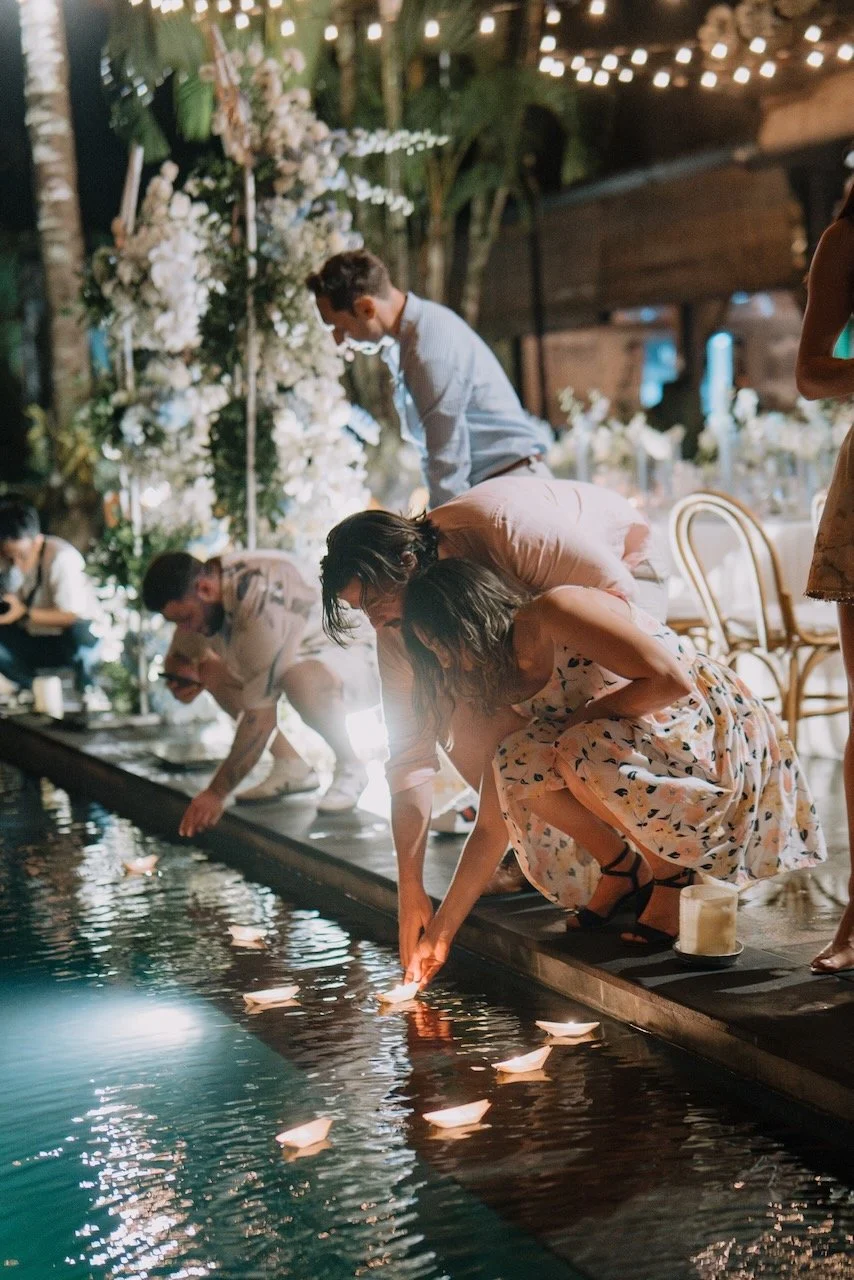 Guests releasing floating candle lanterns into the pool during nighttime Bali wedding celebration under string lights.