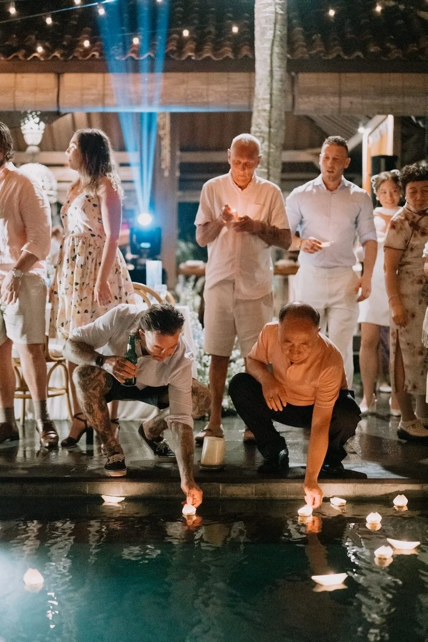Guests releasing floating candles into the pool during nighttime Bali wedding celebration under string lights.