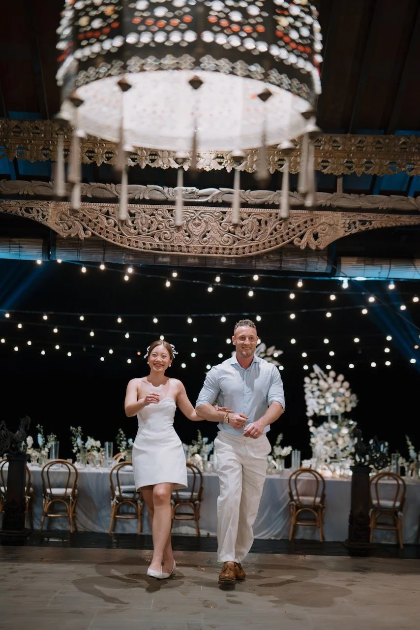 Bride and groom joyfully entering the wedding reception in Bali under ornate lantern and twinkling string lights.