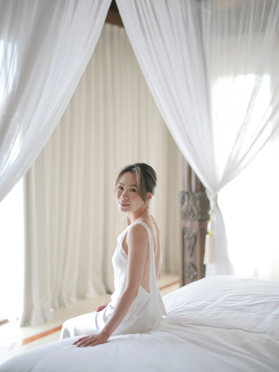 Bride seated on a canopy bed in soft natural light during relaxed Bali wedding morning preparations.