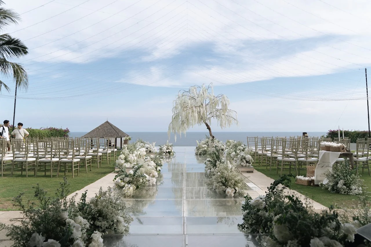 Oceanfront Bali wedding ceremony setup with mirrored aisle, gold chairs and lush white floral arrangements.