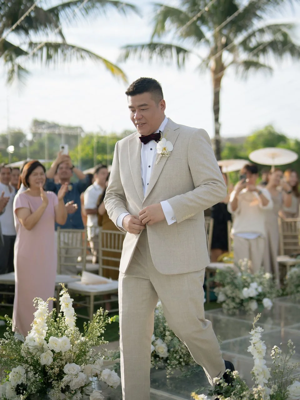 Groom walking down the aisle surrounded by white florals during a tropical Bali destination wedding ceremony.