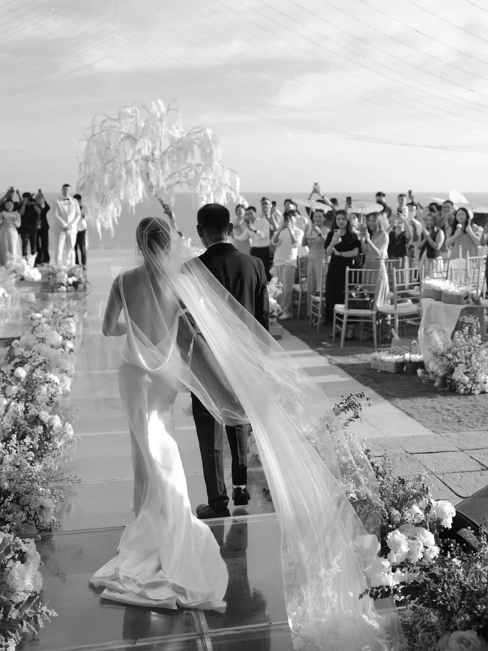 Bride and groom walking hand in hand down a mirrored aisle as guests applaud at an oceanfront Bali wedding.