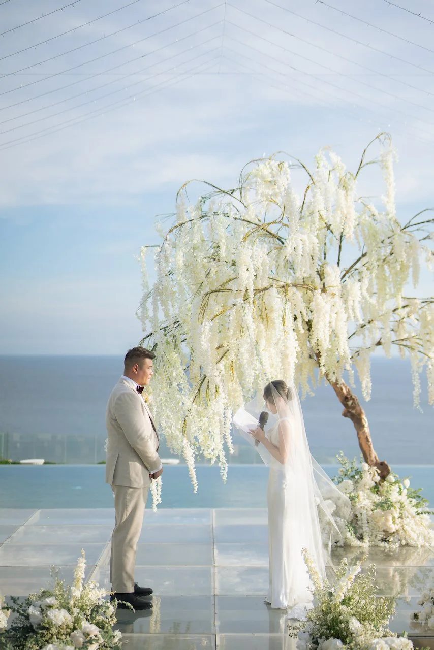 Bride reading vows beneath a cascading white floral installation during a luxury clifftop Bali wedding ceremony.