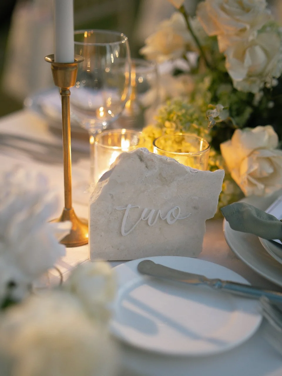 Close up of elegant place setting with stone table number, candles and white flowers at a Bali wedding reception.