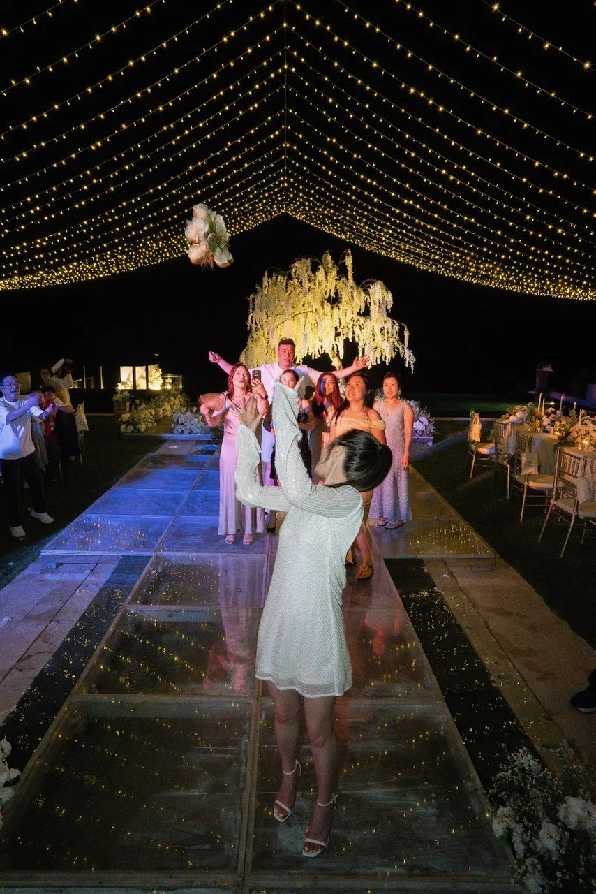 Bride tossing bouquet to guests on a mirrored dance floor under fairy lights at a joyful Bali wedding reception.