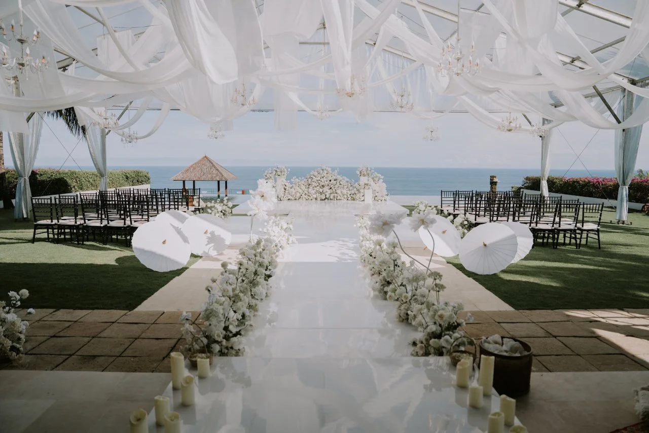 Luxury oceanfront wedding ceremony at The Surga Bali with white floral aisle, draped canopy and chandeliers, planned by Silver Lace Weddings for Sophia and Hamish