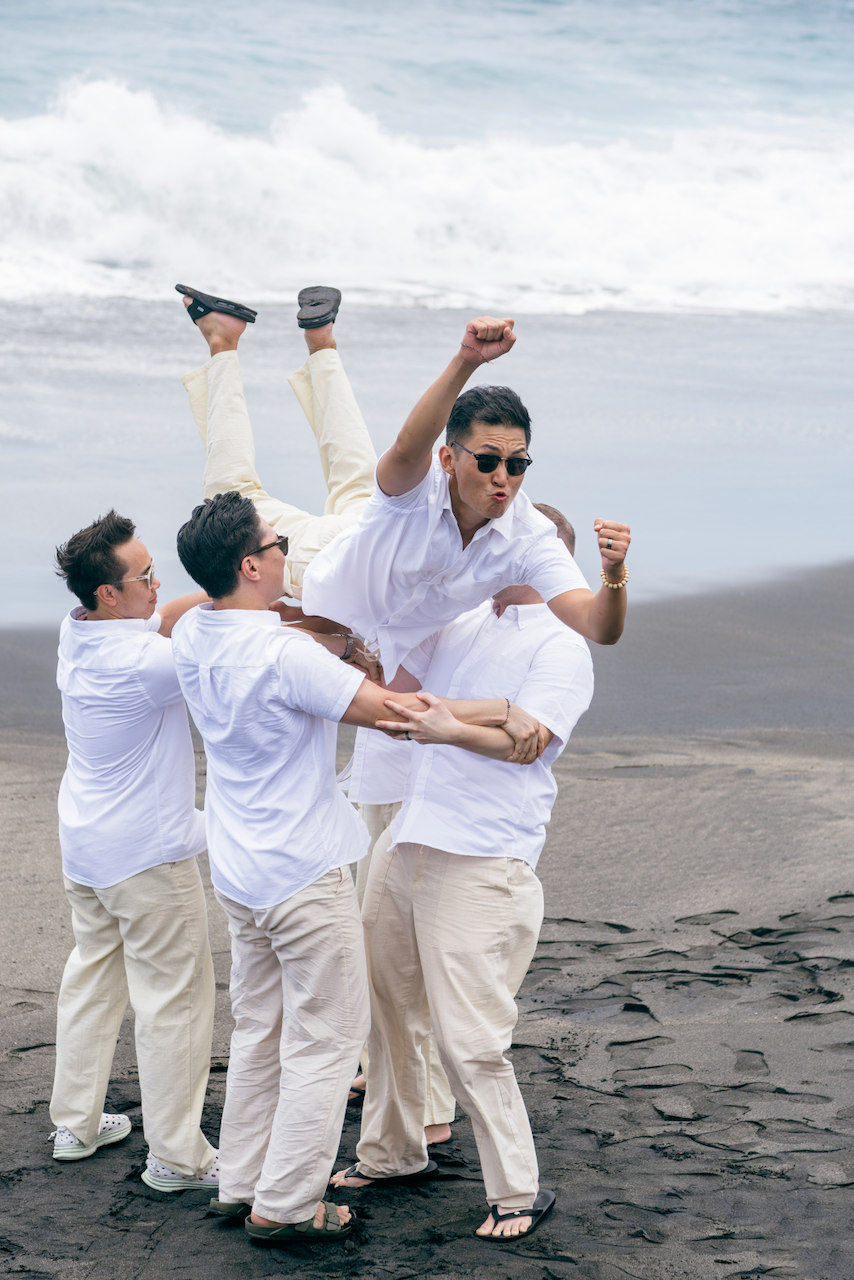 Playful groomsmen lifting the groom on a black sand beach during a Bali destination wedding