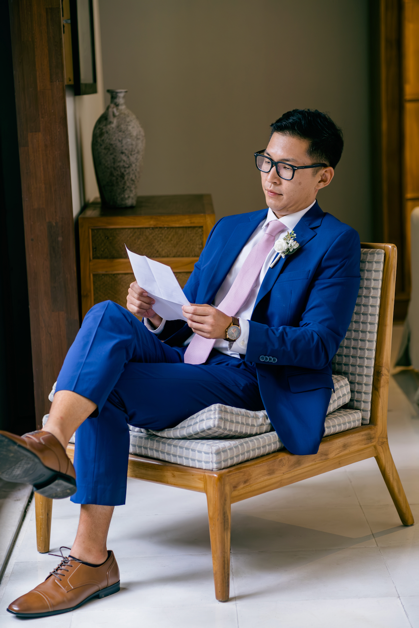 A groom in a sharp blue suit with glasses reads a personal note before his destination wedding in Bali