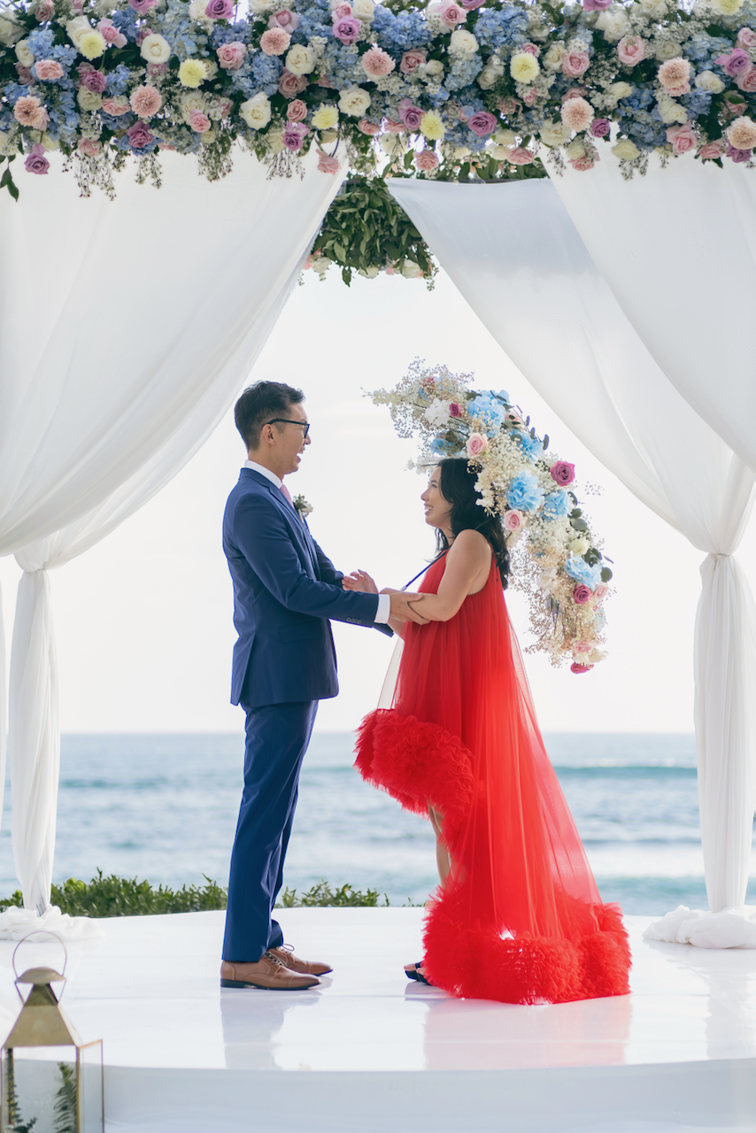 A bride with a custom floral umbrella stands with her groom under a beautifully decorated wedding arch in Bali