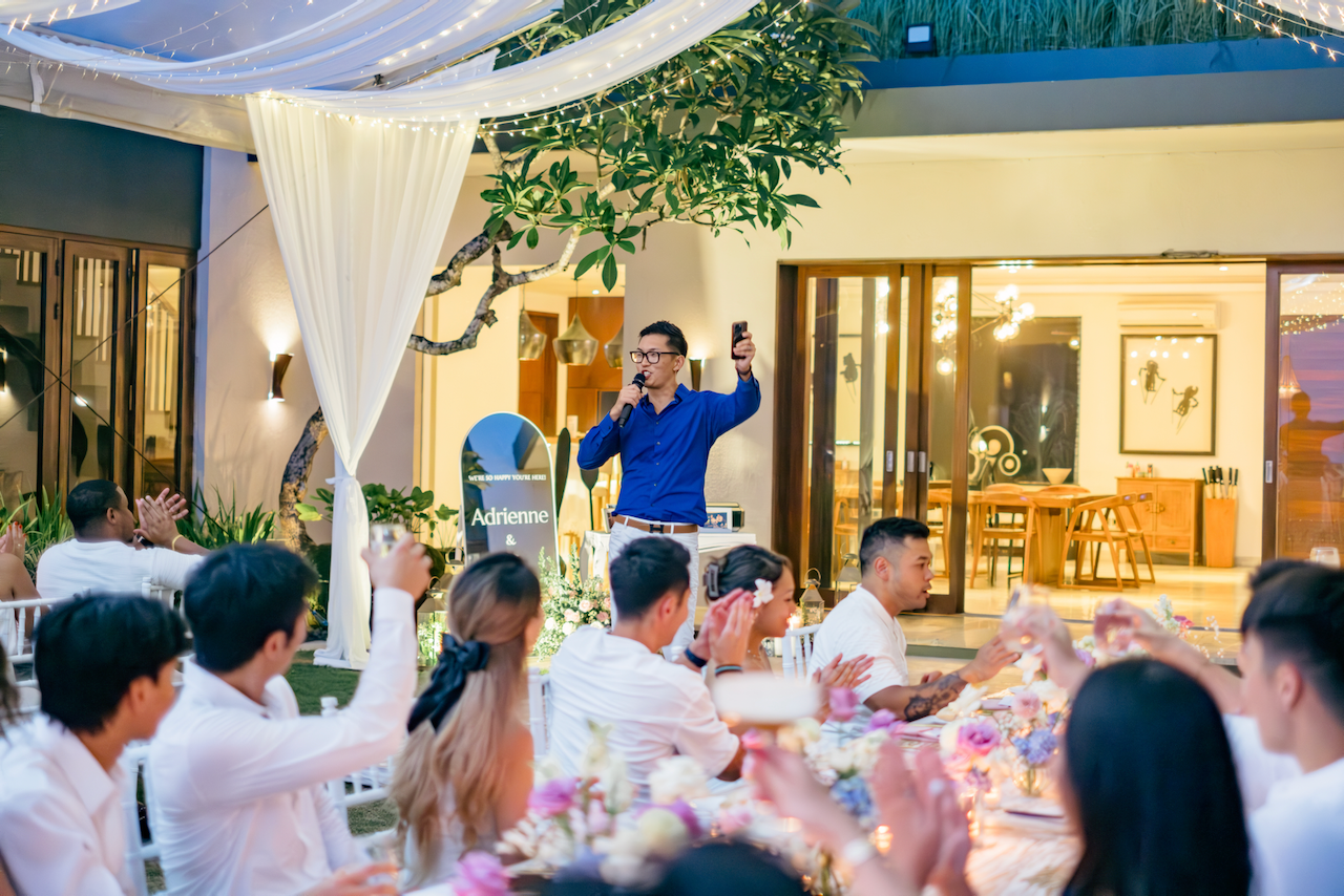 A guest in a blue shirt holds a microphone to give a heartfelt speech at a reception dinner table