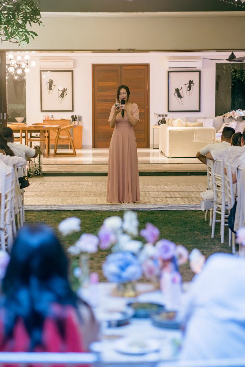 A speaker in a pink gown stands at the reception stage, viewed through a foreground of wedding tables and flowers