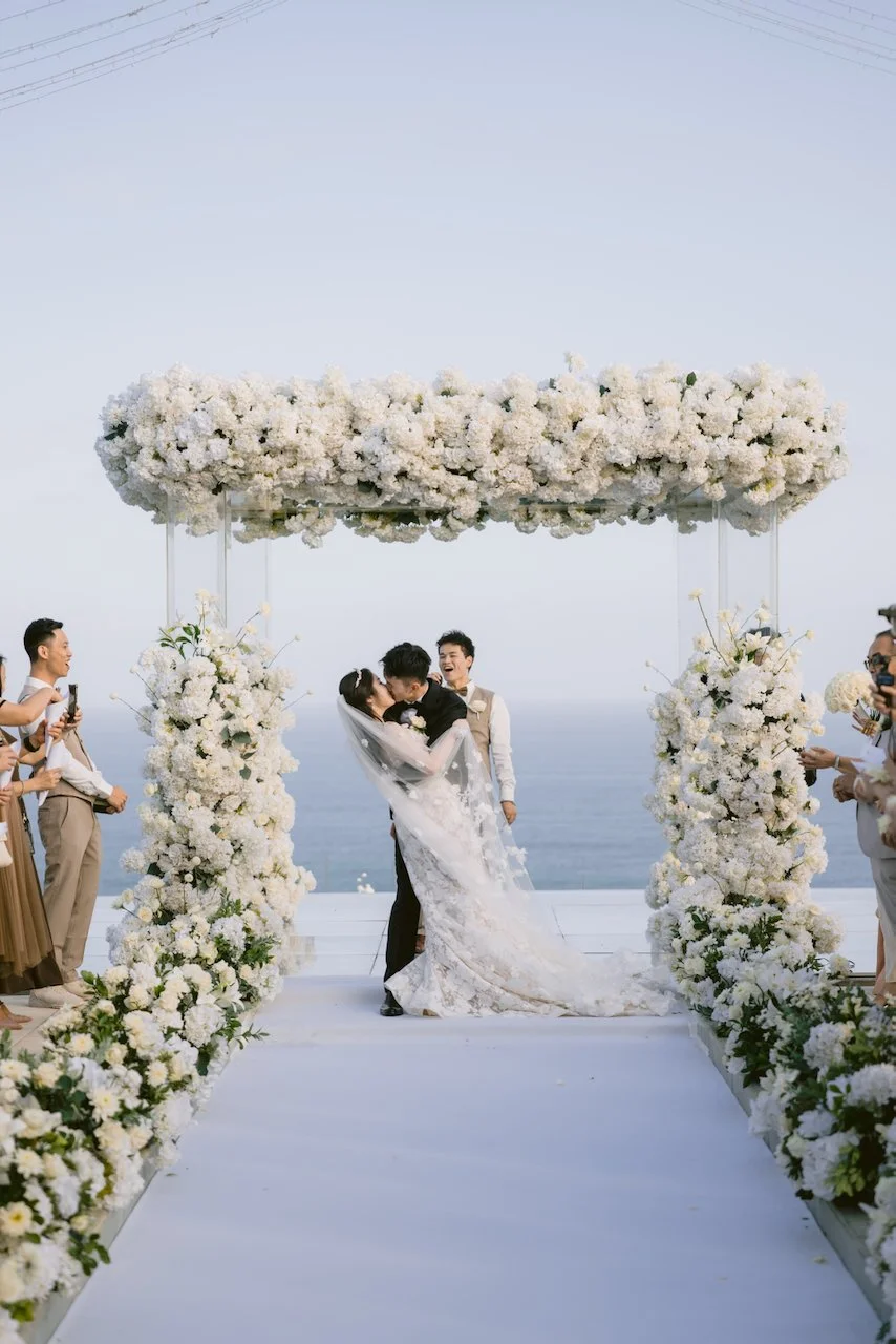 Bride and groom first kiss under floral arch overlooking ocean in Bali