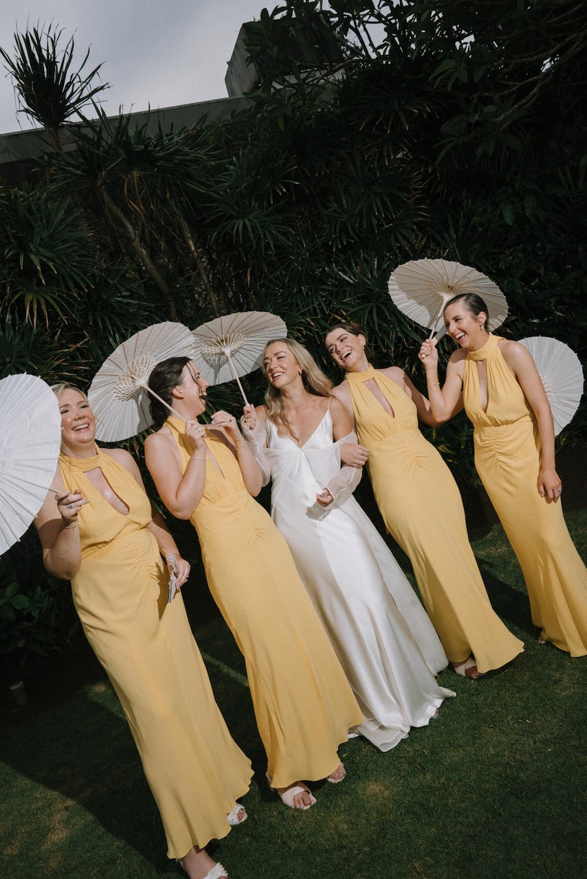 Laughing bride and bridesmaids holding white paper parasols against lush greenery in Seminyak.