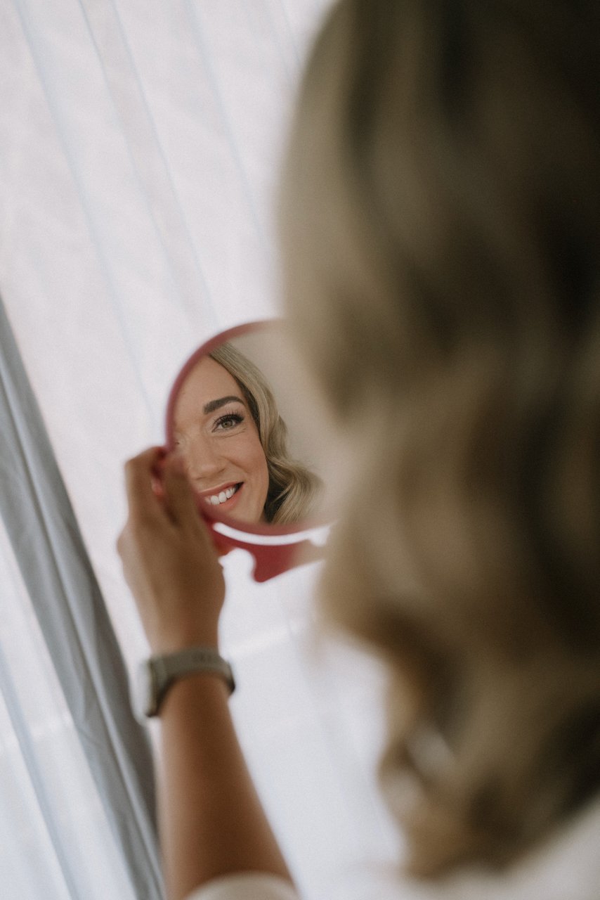 Smiling bride checking her hair and makeup in a pink hand mirror before her coastal destination wedding.