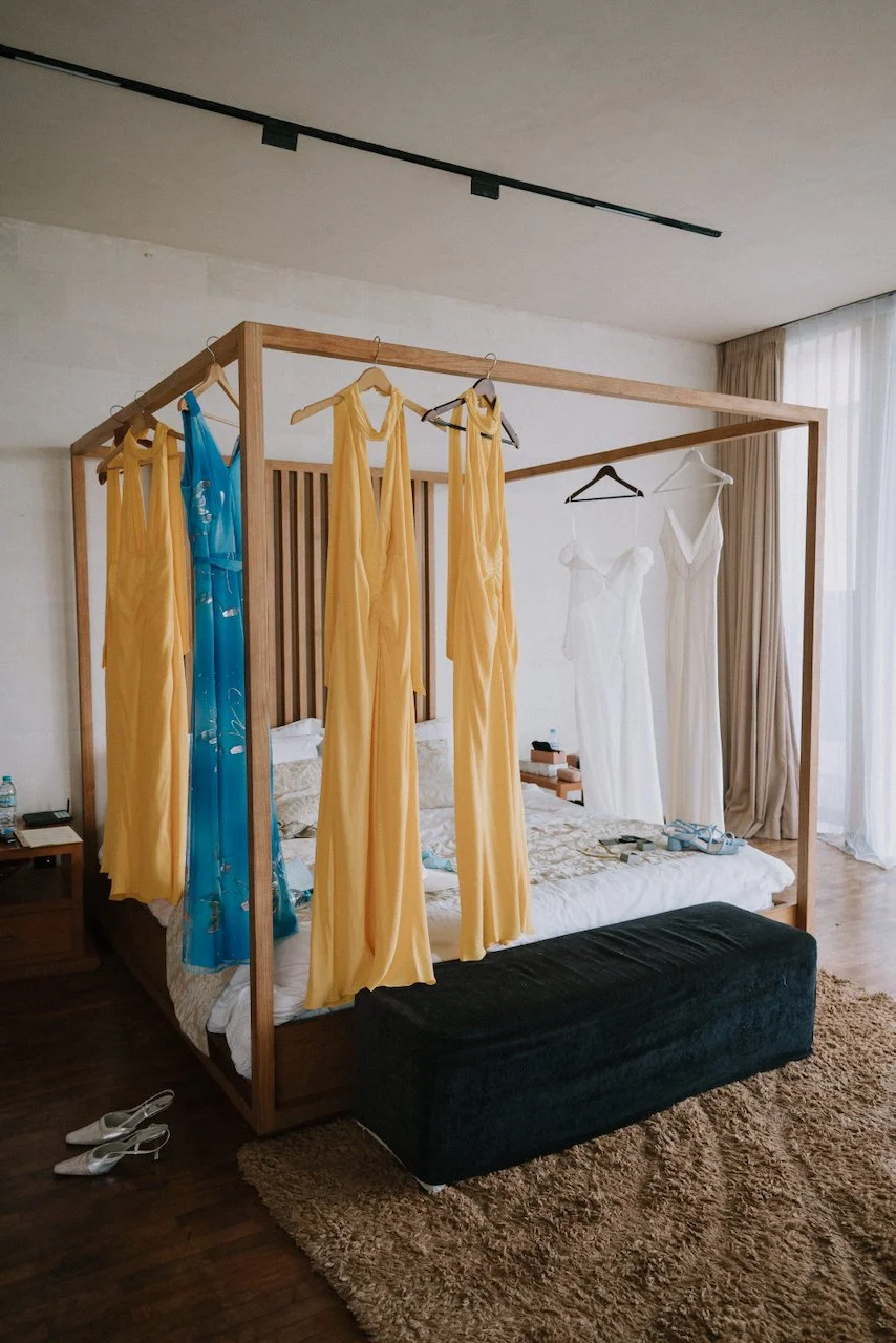 Yellow bridesmaid dresses and a white bridal gown hanging on a wooden canopy bed during Seminyak wedding prep.