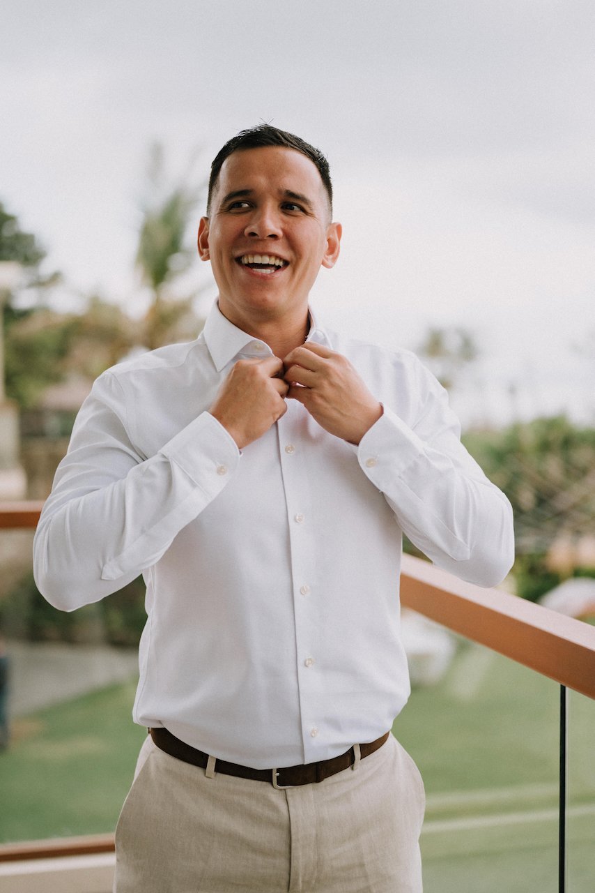 Cheerful groom buttoning his crisp white dress shirt on a bright island balcony.